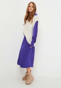 Chaleco de punto crema sobre un vestido de satén morado, que cuenta con mangas largas, un corte holgado y un patrón texturizado. Botas de cordones color marrón claro completan el atuendo.