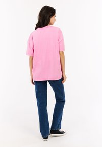 Overmaatse roze katoenen t-shirt met korte mouwen, gecombineerd met blauwe denimspijkerbroeken en zwarte lage sneakers, gezien vanaf de achterkant.