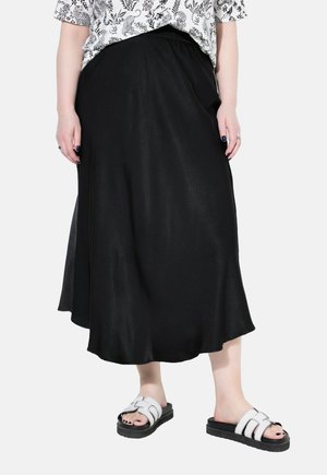 Jupe midi noire, en satin, avec une taille élastique, présentant une silhouette fluide. Associée à des sandales blanches épaisses.