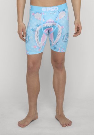 Mann trägt blaue Boxershorts mit weißem und rosa Hai-Kiefer-Grafik sowie PSD-Logo am Bund, steht barfuß auf grauem Boden.