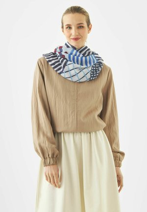 Vrouw met lichtbruine blouse en crèmekleurige rok, met een blauw, wit en kastanjebruin sjaal met patroon, staand tegen een witte achtergrond.