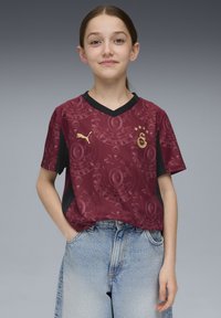 Bordeaux jalkapallo-ottelupaita, jossa on kuvioitu design, mustat yksityiskohdat, V-kaulus ja Puma-logo. Yhdistetty vaaleansinisiin farkkushortseihin.
