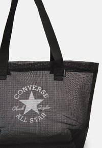 Sac fourre-tout en maille noire avec deux poignées noires. Présente une étoile blanche et le texte "CONVERSE Chuck Taylor ALL STAR" imprimé sur le devant.