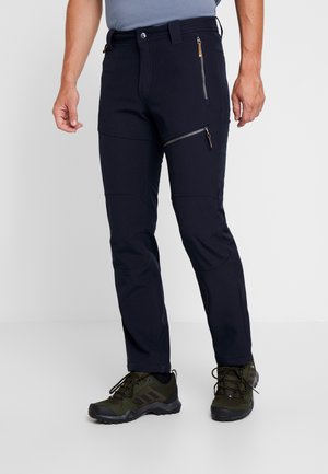 Mężczyzna w granatowych spodniach z zamkniętymi kieszeniami i zielonych butach górskich, stojący na tle białym.