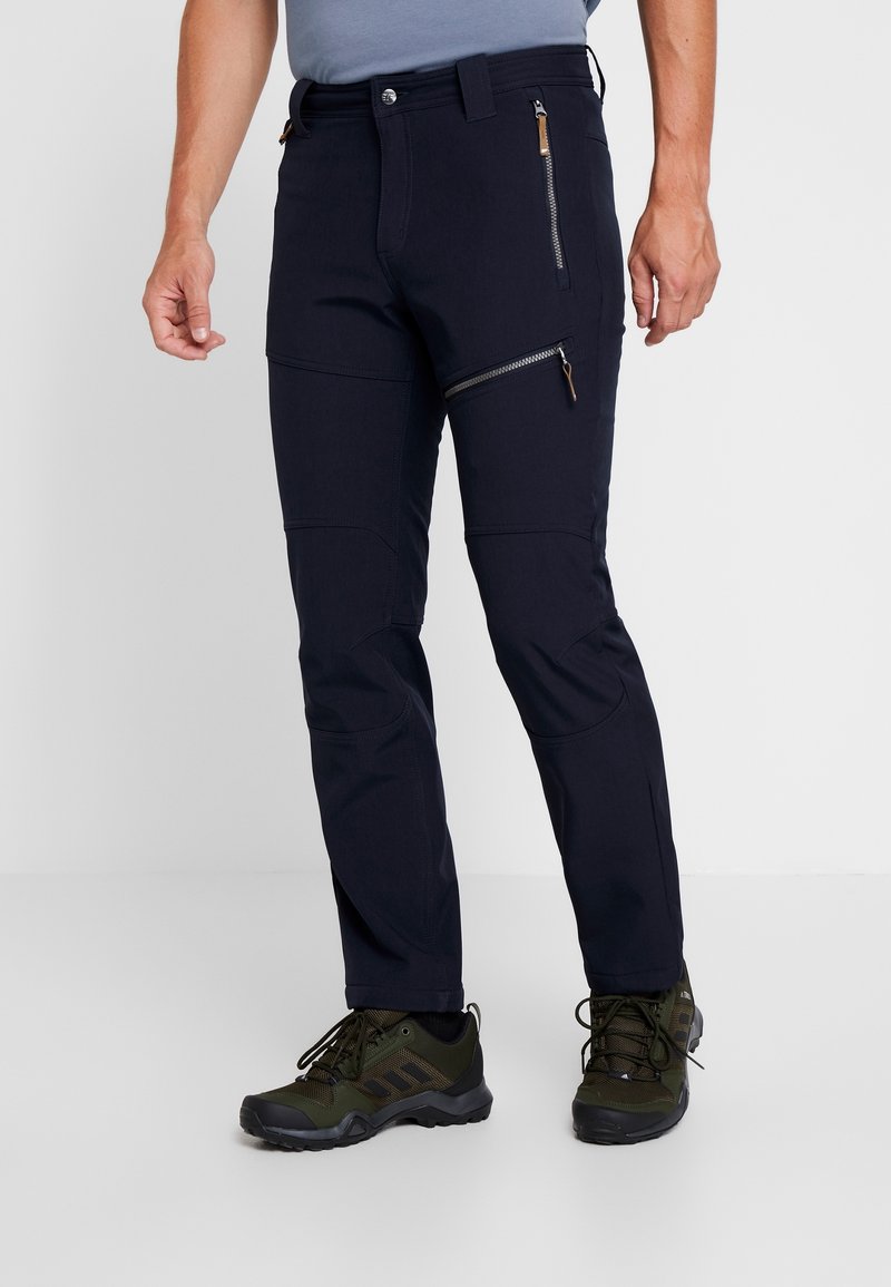 Muž ve tmavě modrých kalhotách se zipovými kapsami a zelených turistických botách, stojící na pozadí z čistě bílého pozadí.