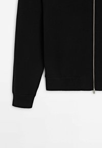 Veste noire à manches longues avec fermeture éclair argentée à l'avant et poignets et ourlet côtelés, montrée de la taille au avant-bras sur fond blanc.