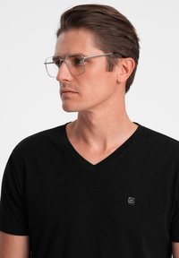 Czarny t-shirt z dekoltem w serek wykonany z bawełny, z małą metką na piersi. Przezroczyste, metaliczne prostokątne oprawki okularów. Krótkie, stylowo ułożone włosy.