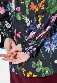 Mani incrociate dietro la schiena, indossando una maglietta nera a maniche lunghe con stampa floreale e di funghi colorati, abbinata a pantaloni rosso scuro.