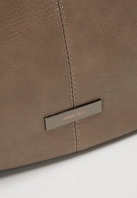 Gros plan sur un matériau en cuir taupe avec une couture verticale et une plaque métallique rectangulaire affichant le texte "ANNA FIELD".