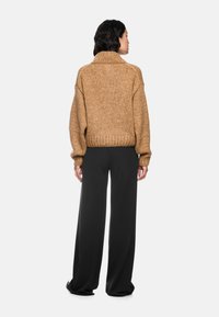 Brauner Strickpullover mit hohem Kragen und geripptem Saum, getragen über schwarzen weiten Hosen. Weiche Textur, entspannte Passform und heruntergesetzte Schulternähte.