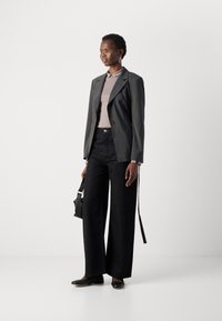 Jean large noir avec des embellissements en argent, blazer gris sur un haut gris clair, et chaussures noires. Le mannequin tient un sac à main noir texturé.