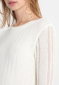 Maglione bianco a maglia con texture a costine verticali e maniche in pizzo trasparente, scollo rotondo e vestibilità ampia. Orecchini a cerchio dorati visibili.