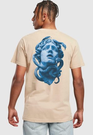Camiseta corta de manga beige con un gráfico azul de una figura drapeada con serpientes alrededor de su cabeza, añadiendo textura y profundidad al diseño.