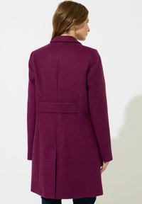Cappotto lungo e aderente in misto lana di un profondo colore viola, con colletto a risvolto, dettagli cuciti sul retro e tasche laterali. Tessuto morbido, lunghezza fino a metà coscia.