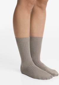 Pair of legs wearing mid-calf, ribbed beige socks, standing on a white surface with a plain white background.