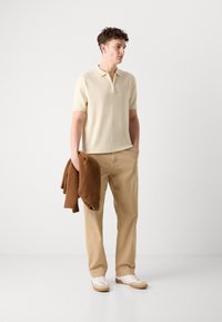 Krämfärgad kortärmad polo med dragkedja, beige byxor, hållande en brun tröja, avslutad med vita sneakers och gummisulor.