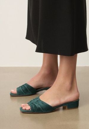 Voeten met groene slip-on sandalen met open neus en lage hak, gecombineerd met zwarte broek met wijde pijpen, staand op een gladde vloer.