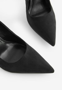 Scarpe con tacco alto in suede nero con punta affusolata e design con ritagli sui lati. Texture liscia, accenti minimalisti e silhouette elegante.