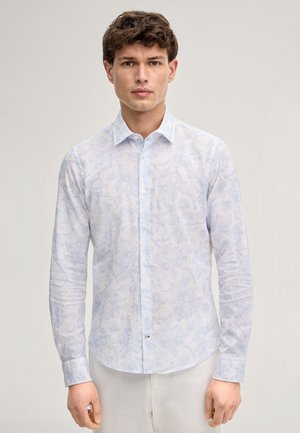 Mann trägt ein hellblaues, langärmliges Hemd mit zartem Blumenmuster und weiße Hosen, steht vor einem einfarbigen Hintergrund.