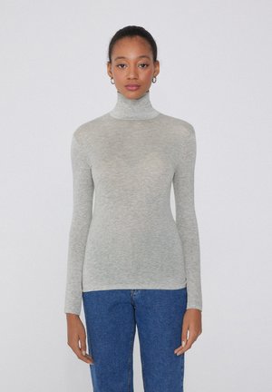 Hellgrauer Rollkragenpullover mit langen Ärmeln, aus weichem, geripptem Stoff. Getragen mit blauen Jeans, die eine figurbetonte Silhouette aufweisen.