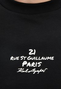 Μαύρο ύφασμα με λευκό κείμενο που γράφει "21 Rue St Guillaume Paris" και μια υπογραφή με καλλιγραφικά από κάτω.