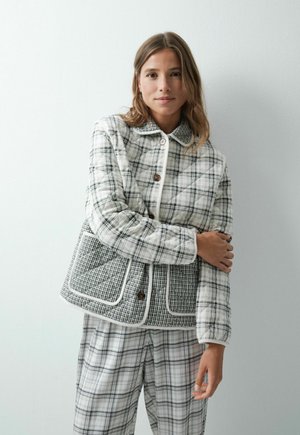 Jeune femme portant une veste matelassée à carreaux blancs et noirs et un pantalon assorti, debout contre un mur blanc uni.