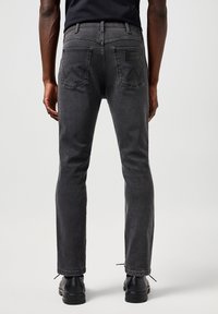 Mann iført svarte smale jeans og svarte sko, stående med ryggen til mot en ensfarget lys bakgrunn.