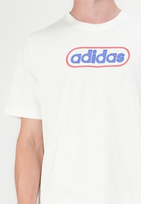 Balts kokvilnas T-krekls ar zilu un sarkanu ovālu logo, kurā ir teksts "adidas" treknajās, izliekta burtiem. Īsas piedurknes un apaļš kakls.