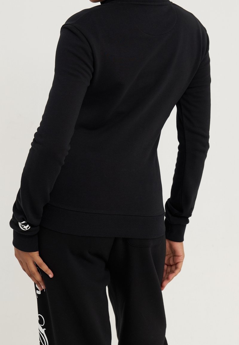 Personne portant un haut noir uni à manches longues et un pantalon noir avec un motif blanc sur le côté, debout dos à la caméra.