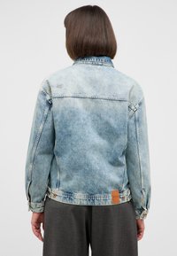 Jasnoniebieska kurtka dżinsowa o lekko wyblakłej fakturze, z kołnierzykiem zapinanym na guziki i skórzaną metką na dolnym skraju. Widok z tyłu ukazuje detale przeszycia.