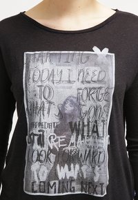 Schwarz langärmliges T-Shirt mit einem grafischen Druck, der gemischten Text und ein Bild in Graustufen zeigt. Weicher Stoff und entspannter Halsauschnitt.