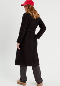 Black, long coat with a textured fabric, belted waist, and long sleeves. Paired with dark trousers and a red cap. Soft, casual look.