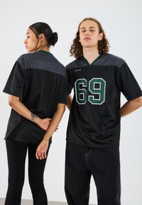 Svart sportsjakke med grønne detaljer, med nummer 69 i stor skrift. Laget av pustende stoff, med kortermede og v-hals.