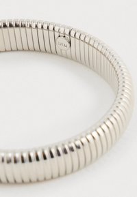 Strieborný kovový náramok s flexibilným, pružinovým dizajnom, ktorý má hladký, lesklý povrch a malú značkovú etiketu.