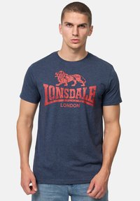 Tengerészeti kék pamut póló, piros oroszlán grafikával és a "LONSDALE" felirattal a "LONDON" felett. Rövid ujjú és kerek nyakú.