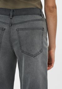 Vue arrière d'une personne portant un jean gris en denim et un haut vert olive, montrant la poche arrière et une partie du bras droit sur fond blanc.