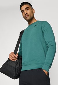 Teal sweatshirt met een ronde halslijn en ribgebreide manchetten, gecombineerd met een zwarte tas met netaccenten. Model houdt de tas op zijn schouder.