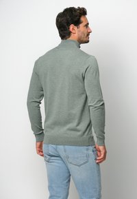 Grauer Strickpullover mit hohem Kragen, taillierter Passform und gerippten Bündchen. Getragen mit hellblauen Jeans, von hinten betrachtet.