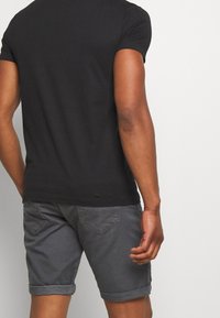 Mężczyzna w ciemnoszarych, podwiniętych szortach i czarnej koszulce z krótkim rękawem, widoczny z tyłu na tle jednolitego białego tła.