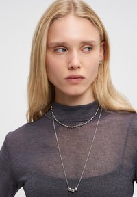 Sølv halskæde med to lag; det ene lag har små, sfæriske detaljer, mens det andet er en delikat kæde. Modellen har en mørk, gennemsigtig bluse på.