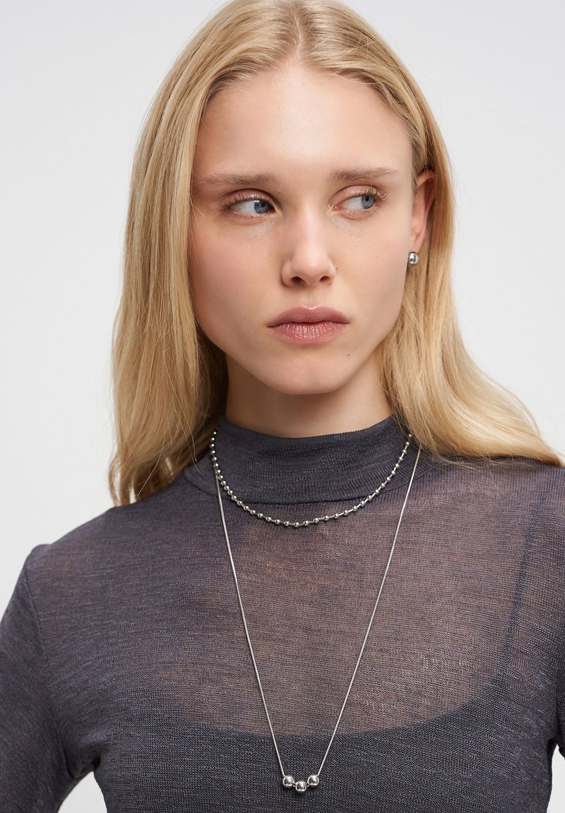 Sølv halskæde med to lag; det ene lag har små, sfæriske detaljer, mens det andet er en delikat kæde. Modellen har en mørk, gennemsigtig bluse på.