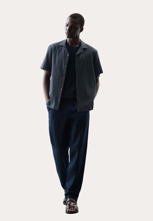 Mężczyzna stojący, ubrany w ciemną koszulę na krótki rękaw z guzikami, dopasowane ciemne spodnie i brązowe sandały, z rękami w kieszeniach.