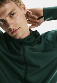 Junger Mann mit hellen Haaren, der eine dunkelgrüne Zip-Jacke trägt und mit den Händen hinter dem Nacken vor einem weißen Hintergrund posiert.