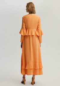 Narancssárga peplum ruha rétegezett fodrokkal, hosszú ujjakkal és hátsó cipzáras záródással. Simaságot tükröző szövet, bokáig érő hosszúság, magas sarkú szandálokkal párosítva.
