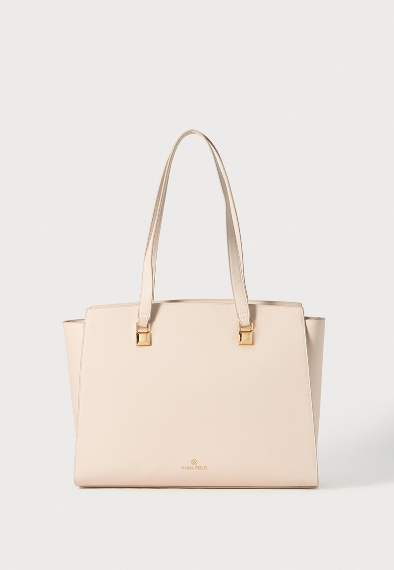 Beige tote taske med struktureret form, syntetisk materiale, to lange håndtag, hardware i guldfarve og logo i bunden midt.