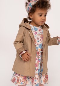 Jasno beżowa kurtka z kapturem i zapięciem na zamek. Pod nią kwiecista sukienka w delikatnych pastelowych kolorach, z falbanami na mankietach i u dołu.