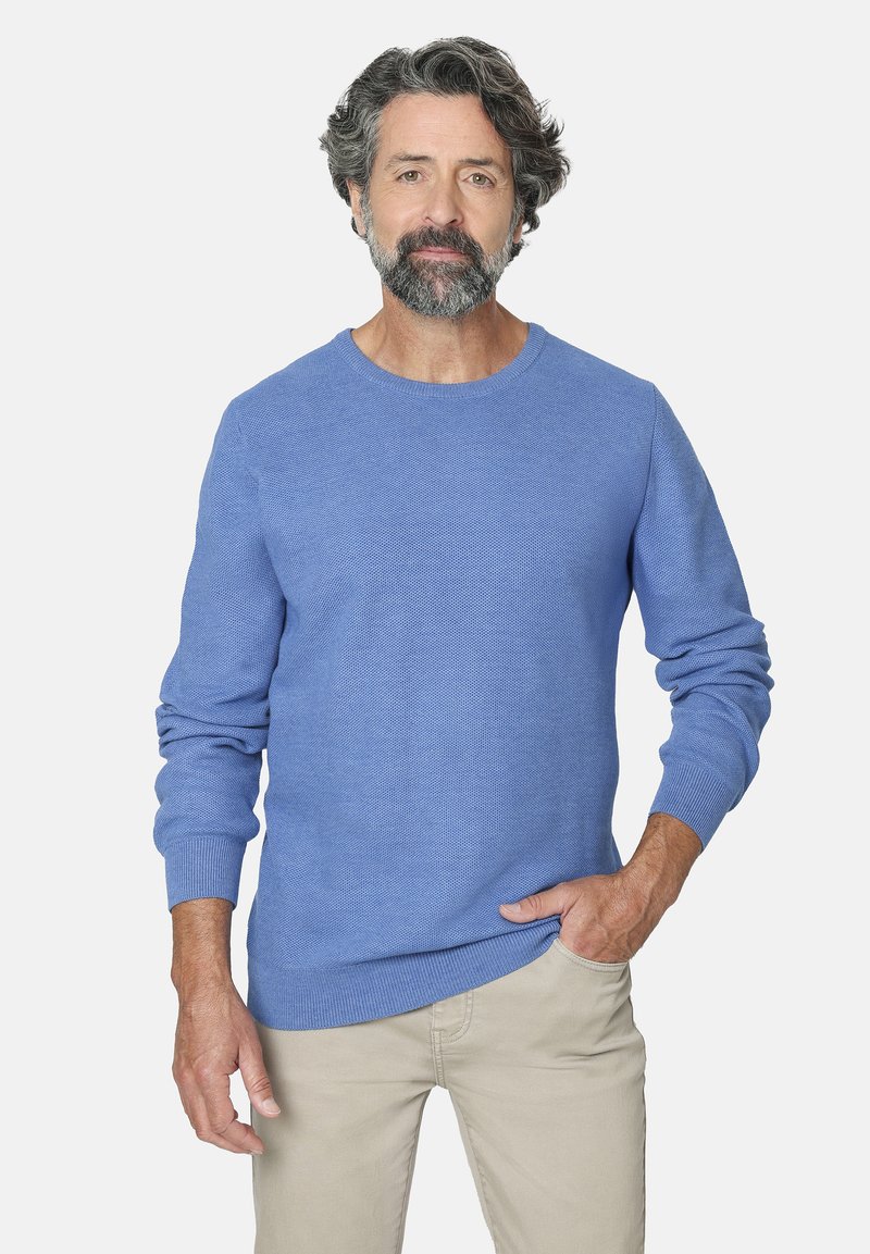 Hombre de mediana edad con cabello y barba canosos, vistiendo un suéter azul claro y pantalones beiges, de pie con una mano en el bolsillo.