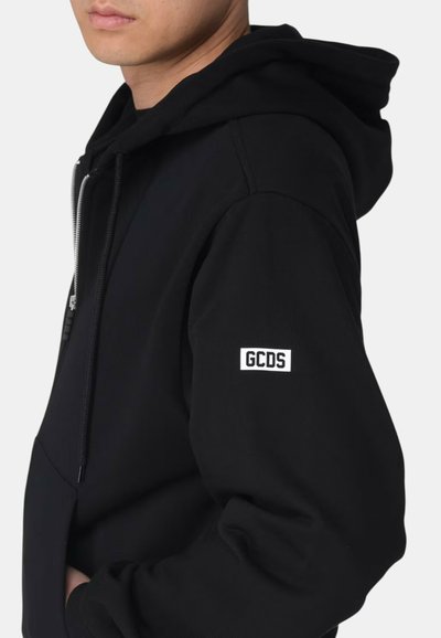 Mężczyzna w czarnej bluzie z kapturem zapinanej na suwak z naszywką z logo GCDS na lewym rękawie, ręce w przedniej kieszeni, na jednolitym jasnym tle.