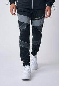 Pantalones deportivos negros con acentos grises y un diseño geométrico, fabricados con material ligero, combinados con zapatillas blancas.