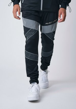 Pantaloni sportivi neri con dettagli grigi e un design geometrico, realizzati in materiale leggero, abbinati a sneakers bianche.